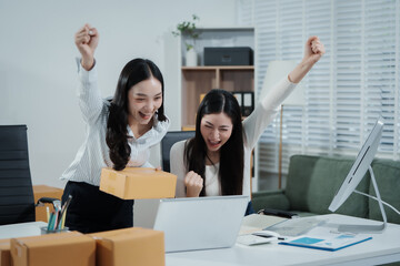 Two Asian female entrepreneurs celebrate a major online sales achievement,cheering happily while checking their laptop in an e-commerce office surrounded shipping boxes,excitement,business success