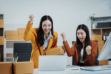 Two Asian female entrepreneurs celebrate a major online sales achievement,cheering happily while checking their laptop in an e-commerce office surrounded shipping boxes,excitement,business success