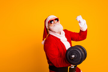 Santa in red suit lifts dumbbell on bright yellow background with shades and festive vibe for christmas promotion and fitness holiday theme