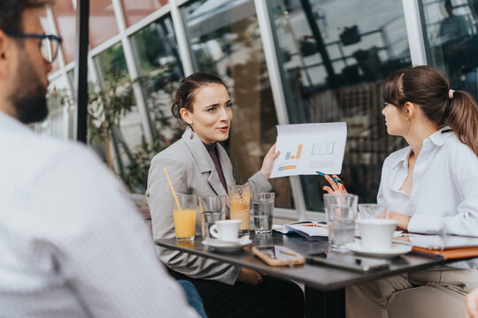 A group of colleagues collaborating over graphs and charts in an open cafe environment, suggesting teamwork, business strategies, and creative communication over coffee and refreshing drinks.
