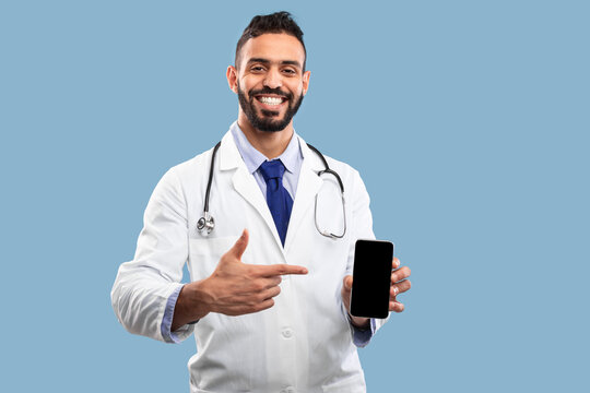 A healthcare professional proudly points at his smartphone, showcasing a friendly demeanor while wearing a lab coat and stethoscope. The background is a solid color.