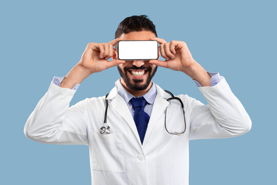 A cheerful doctor wears a lab coat and stethoscope, holding a smartphone in front of his face. The background is a solid light blue, creating a vibrant and engaging atmosphere. - Powered by Adobe