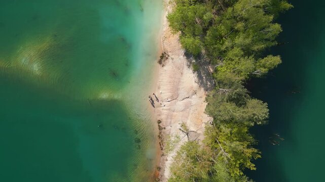 A bright sandy and rocky peninsula extends into turquoise water, surrounded by dense forest and clear underwater structures beneath the calm surface.