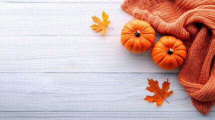 Two small orange pumpkins and two autumn leaves are arranged on a white wooden surface next to a cozy orange knitted scarf. The scene is lit with soft light, cr