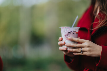 A woman in a bright red coat holds a creamy berry smoothie in a clear cup with a straw outdoors. The focus is on her hands and a sparkling engagement ring, conveying casual, relaxed refreshment.