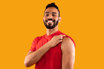 A young man happily shows his arm with a bandage after receiving a vaccination. He stands in front of a vibrant orange background, exuding positivity and confidence.