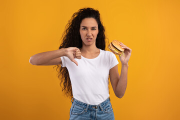 A woman with curly hair stands against a yellow background holding a burger and showing a thumbs...