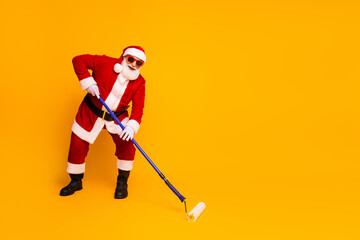 Obraz premium Santa in red suit paints holiday vibes with roller yellow background capturing merry season spirit and playful home style for festive stock photo