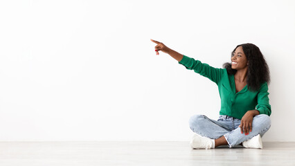 A woman with curly hair sits cross-legged on the floor, wearing a green top and jeans. She points...