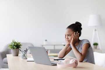 Fototapeta premium An afro american girl takes a break from her online lessons, looking thoughtfully at her laptop in a cozy living room. She appears tired and bored, reflecting the new normal of remote learning.