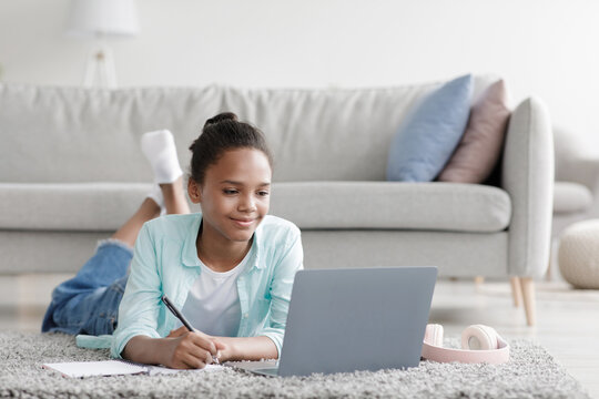 Adolescent girl lies on the floor in her living room, studying and engaged in an online lesson on her laptop. She takes notes while smiling and enjoying the learning process.