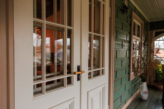 Close up of cream double doors with rectangular glass panes, brass handle, and candle sconce on green clapboard siding, with potted plants and a white floral pitcher