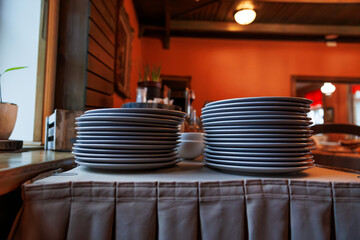 Two tall stacks of white plates sit on a skirted buffet by a window, separated by a small bowl. Warm pendant lights glow, with chairs and tableware beyond.