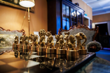 A row of gold chess pieces stands on a reflective glass board in an opulent sitting room, with carved wood furniture and a glass cabinet, warm evening light.
