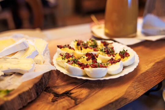 Halved deviled eggs sit on a white plate with bacon and microgreens on a wooden board beside soft rind cheese in warm indoor evening light, styled for casual tasting.