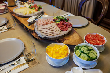 Sliced ham, roast pork, salami, and herbs rest on round wood boards with bowls of cucumber, tomato, and yellow pepper, plates and glasses set for a shared meal.