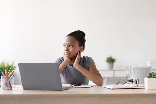 A teenage girl sits at a table with a laptop, looking thoughtfully into space. She navigates online lessons in her living room, adapting to the new normal during the covid-19 pandemic.