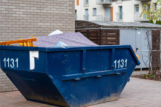 Blue construction waste container with number placed by the housing wall filled with materials covered with protective felt