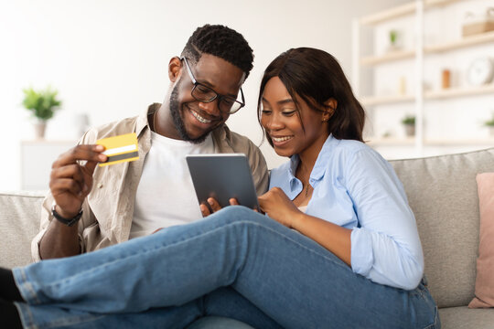 A black couple relaxes on their couch, using a tablet and credit card to shop online. They are engaged and happy while browsing for items during the weekend at home.