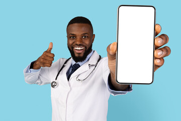 Joyful Black Doctor Demonstrating Blank Smartphone And Showing Thumb Up At Camera, Happy African American Male Therapist Recommending Mobile App Or Website While Posing Over Blue Background, Mockup