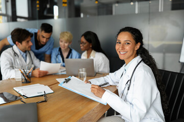 Attractive young woman doctor working on report while having morning meeting with international colleagues, firendly professional team discussing clinical cases, using laptop, having training