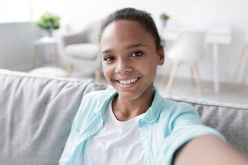 A cheerful teen girl with a beautiful smile is taking a selfie while sitting on a couch in her living room. She is engaged in an online meeting during the covid-19 quarantine period.