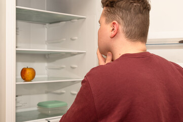 Young man looking into an almost empty refrigerator with only a single apple inside, symbolizing financial hardship, rising food prices, and the struggle to afford groceries during an economic crisis.