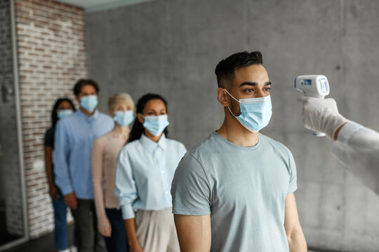 Multiracial men and women in protective face masks checking body temperature before vaccination against coronavirus at clinic, unrecognizable nurse hand with electronic thermometer