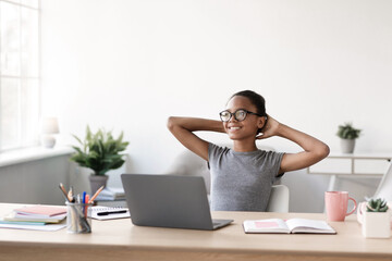 A teenage black girl in glasses relaxes at her table while looking at the space in front of her. She enjoys a break from studying at home in a bright living room.