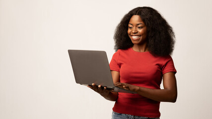 A cheerful African American woman stands in front of a white background while holding her laptop. She radiates happiness as she engages in remote work, showcasing the joy of freelancing.