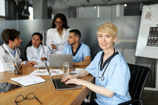 Attractive blonde lady doctor typing on laptop keyboard while having morning breefing with multiracial colleagues, firendly professional team discussing clinical cases, having training - Powered by Adobe