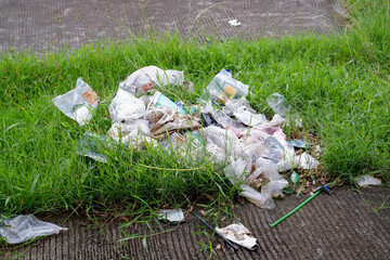 A pile of disposable plastic bags, bottles, and other trash sits in overgrown grass along a sidewalk. This image highlights litter, waste, and environmental neglect in urban outdoor settings.
