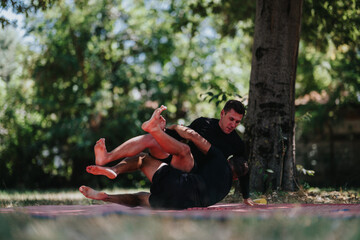 Two men in black shirts grapple on a red mat under a tree in a sunlit park, showcasing fitness, strength, and competitive wrestling outdoors. Ideal for sports, training, and martial arts imagery.