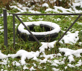 A snow-covered old used car tire lies on the grass near a fence