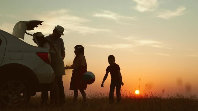 Playful family at sunset by car. cheerful children parents going on travel road trip by automobile packing baggage putting luggage in trunk kids playing ball. Packing preparation for family picnic.