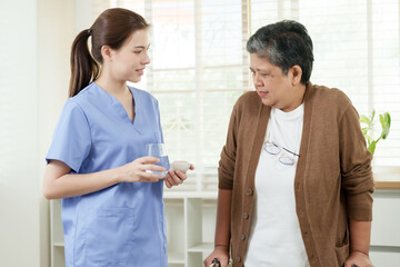 Obraz premium Asian senior woman receiving glass of water and medicine from female physiotherapist during home therapy treatment routine showing caring relationship and physical support recovery