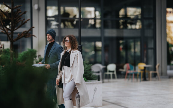 Two fashionable professionals walk outside a glass-front office, carrying documents with a confident, urban vibe and contemporary style.