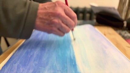 close up side view of man painting canvas of blue sea and sky with paint brush