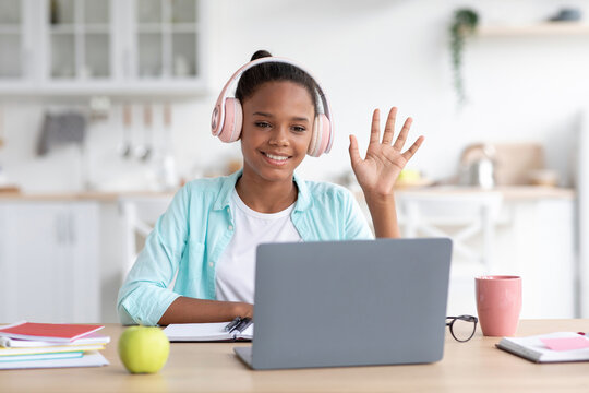 A thoughtful adolescent African American girl is studying at home. She is using a laptop for online classes, smiling, and waving her hand while wearing headphones.