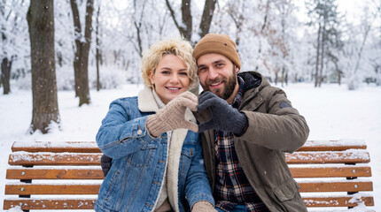 Romantic winter couple forming a heart shape together on a snowy park bench.