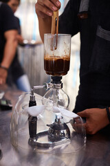 Bali, Indonesia, November 18, 2015. Close up view of man preparing coffee using siphon coffee maker, stirring grounds with wooden stick, focusing on brewing process in modern cafe