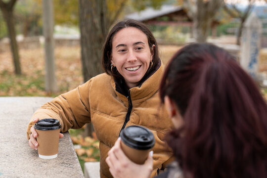 Young women friends enjoying a relaxed conversation outdoors, drinking coffee in a park during autumn - Powered by Adobe