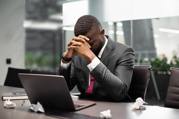 Upset african american man in suit manager sitting at table in front of laptop, leaning on his hands, hiding face, looking for creative solutions for business, need for novel ideas, copy space