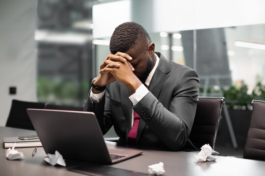 Upset african american man in suit manager sitting at table in front of laptop, leaning on his hands, hiding face, looking for creative solutions for business, need for novel ideas, copy space - Powered by Adobe
