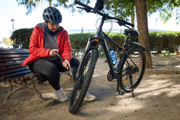 Woman sitting on bench inflating mountain bike tire using a portable hand pump in a park.