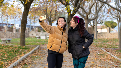 Happy young women taking selfie with smartphone in autumn park, enjoying friendship and togetherness outdoors