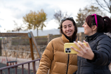 Happy young women friends taking selfie with smartphone outdoors in park, sharing cheerful moments in autumn