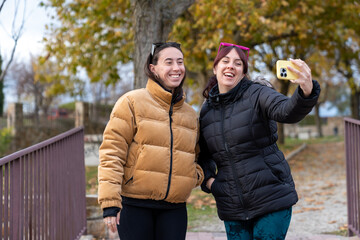 Two jovial women giggling as they take a selfie on a smartphone, dressed in cozy jackets on a crisp autumn day