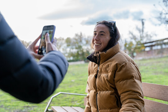 Woman smiling while friend takes smartphone picture in park, enjoying social media memory capture - Powered by Adobe