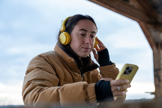 Woman listening to music with yellow headphones, holding a smartphone outdoors, connecting with technology and digital lifestyle